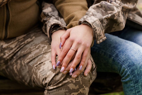 A close-up image showcasing hands clasped together, one wearing a military camouflage jacket and the other denim jeans, with a diamond ring visible, suggesting love, commitment, and partnership