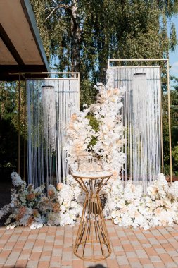 Çok şık bir açık hava düğün töreninde parıldayan beyaz perdelerle süslenmiş altın geometrik bir kemer ve renkli şeftali ve krema çiçekleri sergilenir.