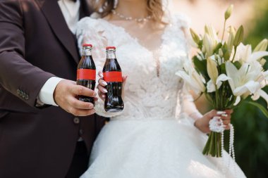 Yeni evli bir çift zarif bir düğün elbisesi içinde ellerinde beyaz zambaklar olan klasik Coca-Cola şişeleriyle neşeli bir kadeh kaldırıyor. Aşkı, kutlamayı ve modern aşkı sembolize eder.