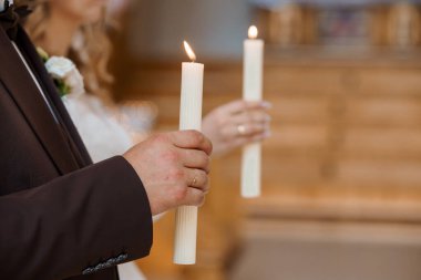 Bir kilise töreninde damat ve gelinin ellerinde yanan beyaz mumlar olduğu samimi bir düğün anı. Yakın çekim romantizm, inanç ve gelenek taşır.