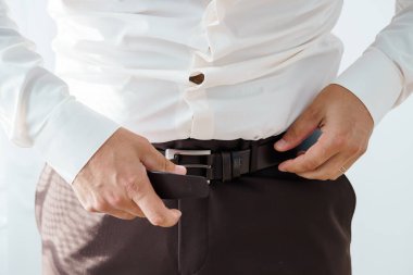 Yakın plan çekimde, beline koyu deri kemer takmış kahverengi pantolon ve açık renk gömlek giyen bir adam resmedilmiş.