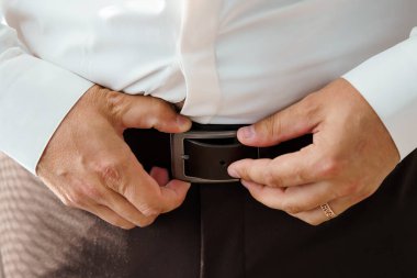 Yakın plan çekimde, düğün hazırlıkları sırasında kahverengi deri kemerini siyah bir tokayla bağlayan bir damat görülüyor. Ellere, resmi giysilere ve samimi anlara odaklan.