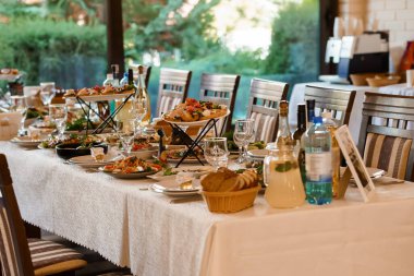 Gourmet aperatifleri, şarap şişeleri, cam kaplar ve ekmek sepetleriyle dolu, parlak, modern, yemyeşil bir restorana bakan bir düğün masası. Sevinçli bir vesile sunar