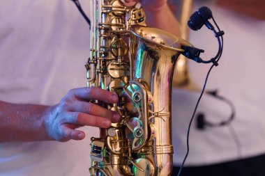 Canlı olarak çalınan pirinç saksafonun detaylı bir görüntüsü, yakınlarda bir lavalier mikrofon, renkli sahne ışıkları altında müzik performansının özünü yakalıyor.