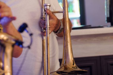 Altından bir trombon çalan, slaytı ayarlayan ellere odaklanan, beyaz tişört ve saat giyen, kapalı mekanda hafif ışıklandırılmış arka planı bulanık bir müzik mekanında