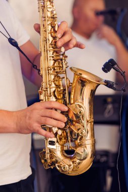 Canlı performans sırasında çalınan altın bir alto saksafonun yakın çekimi, yanında beyaz tişörtlü ve mikrofonlu bir müzisyen, canlı bir müzik olayının enerjisini yakalıyor.