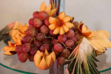 Kırmızı üzümlü renkli bir meyve tabağı, oyulmuş şeftali çiçekleri, ve cam masaya yerleştirilmiş ananas dilimleri, yemek servisi, partiler veya sağlıklı yaşam tarzı görselleri için ideal.