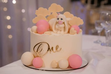 Güzel dekore edilmiş bir bebeğin ilk doğum günü pastasında pembe bir kalp taşıyan sevimli beyaz bir tavşan, pastel renkli balonlar, bulutlar ve yumuşak krema tabanlı altın harflerle çevrili.