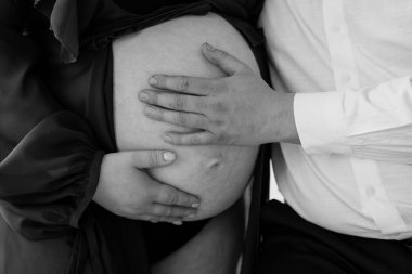 Hamile bir kadının göbeğinin yumuşak siyah ve beyaz portresi. Elleri nazikçe sarkıyor. Hamilelik boyunca samimiyet, beklenti ve annelik bağlarını yakalıyor.