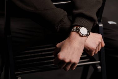 Siyah süveter giyen şık bir adam siyah bir sandalyede oturan parlak gümüş bir saat sergiliyor. Modern zarafeti ve minimalist modayı ön plana çıkarıyor.