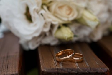 Yumuşak beyaz bir gül buketinin yanında duran iki altın alyansın yakın çekimi aşk ve bağlılığı sembolize ediyor, düğün ya da nişan teması için mükemmel bir ortam.