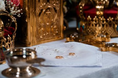 İki altın alyans, süslemeli altın dini eserlerin yanında beyaz dantel bir kumaşın üzerinde duruyor. Çiçek aranjmanları ve sıcak ışıklandırmalarla kurulmuş Doğu Ortodoks kilisesinde ikonlar var.