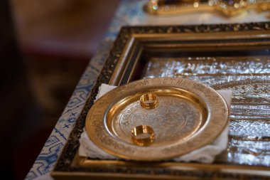 Zengin mavi ve altın seremoni teknikleriyle çevrili dini bir düğün töreninde, karmaşık bir şekilde işlenmiş altın bir tabağa yerleştirilmiş iki altın alyansın yakın çekimi.