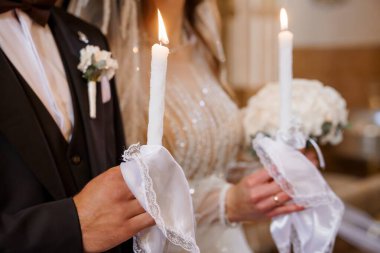 Gelin ve damadın düğün törenlerinde beyaz dantele sarılı mumları tuttukları yakın plan, ılık altın renkleri ve zarif kıyafetleriyle hafif aydınlatılmış bir kilise ortamında birlik ve aşkı simgeliyor.