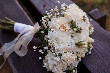Bebek nefesiyle süslenmiş romantik, beyaz bir düğün buketi ve güller kırsal bir ahşap bankta duruyor. Çiçeklerin arasına yerleştirilmiş altın alyanslar, aşkı ve bağlılığı çağrıştırıyor.