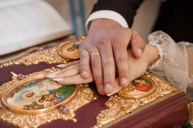 Bir düğün töreninde zengin dekore edilmiş bir dini kitaba sıkıca sarılmış bir erkek ve kadın eli. Altın rengi detaylar, ikonlar ve beyaz danteller sergileniyor. Birlik ve geleneği simgeliyor.