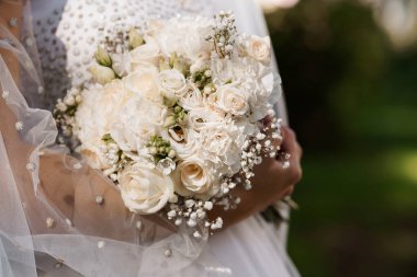 Üzerinde bebek nefesi olan beyaz bir gül buketi tutan bir gelin resmi. Detaylı dantelli bir gelinlik ve duvak takıyor. Yumuşak doğal ışıkta, bulanık yeşil bir bahçe arka planıyla, romantizm ve zarafeti çağrıştırıyor.