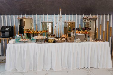 Taze meyve, hamur işi ve tatlılardan oluşan gösterişli bir açık büfe. Beyaz örtülü masa üzerinde altın aksanlar ve mumlar modern bir etkinlik alanında sergileniyor.