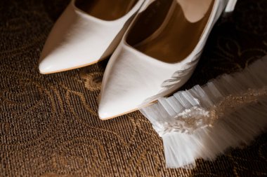 Nazik bir tül jartiyer ile eşleştirilmiş zarif, beyaz uçlu gelinlik ayakkabıları, boncuklarla süslenmiş, desenli kahverengi bir halı üzerinde dinlenen, rafine edilmiş bir düğün hazırlığı anını yakalıyor