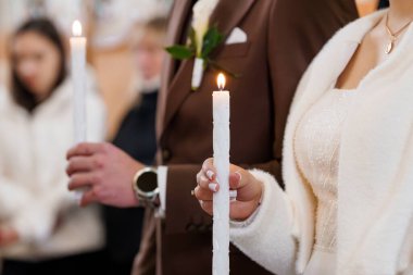 Düğün törenlerinde uzun beyaz mumlar tutan bir gelin ve damadın yakın çekimi. Bir kilisenin ayininde yumuşak ortam ışığı ve çiçek aksanıyla çevrili.