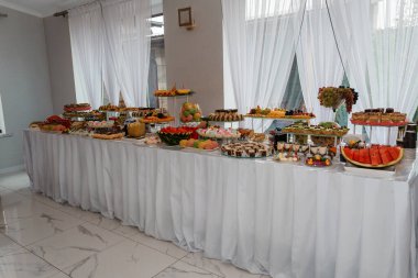 Kekler, pastalar, taze meyveler ve renkli ikramlarla dolu güzel bir tatlı büfesi. Uzun beyaz bir masa üzerinde, parlak kapalı bir alanda, saf perdelerin altında.