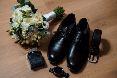 Beyaz gülleri ve yeşillikleri olan güzel bir düğün buketi, ahşap bir yüzeyde siyah ayakkabı, kemer, saat ve parfüm şişesinin yanında oturur.