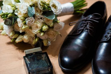 Beyaz gül buketi, siyah deri ayakkabılar ve ahşap bir masanın üzerinde parlak bir parfüm şişesi içeren romantik bir düğün düzenlemesi. Gelinlik hazırlığının ve sofistikeliğin özünü yansıtıyor.