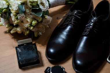 Damadın düğün hazırlıklarının sofistike bir düzenlemesi. Parlak siyah deri ayakkabılar, parlak bir parfüm şişesi, kol saati ve yumuşak doğal ışıklandırmayla sıcak ahşap bir masaya kurulmuş narin bir çiçek buketi.