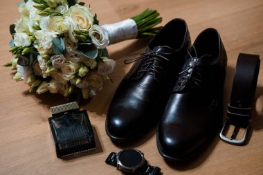 Siyah deri ayakkabılar, uyumlu bir kemer, kol saati, kolonya şişesi ve sıcak ahşap bir yüzey üzerinde bir buket beyaz gül içeren şık bir düğün hazırlığı.