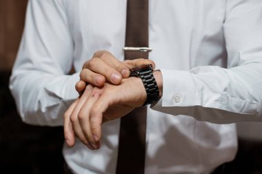 Resmi giyinmiş bir adam, parlak siyah saatini kontrol ediyor. Keskin beyaz gömlek ve siyah kravat takarak hassasiyet ve profesyonelliği vurguluyor.