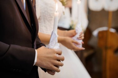 Gelin ve damadın düğün törenlerinde mum tutarken çekilmiş yakın plan bir fotoğraf. Samimi el ele tutuşmalar, zarif kıyafetler ve kilise ortamında hafif bir aydınlatma.