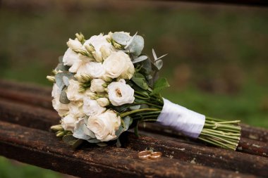 Güller ve okaliptüs yapraklı romantik beyaz bir gelin buketi eskimiş ahşap bir bankta duruyor, evlilik yüzükleriyle birlikte, zamansız aşkı ve doğal zarafeti çağrıştırıyor.