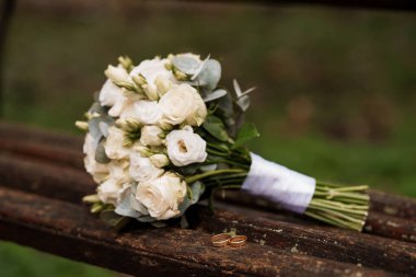 Güzel düzenlenmiş okaliptüs yapraklı beyaz bir gül buketi, yıpranmış ahşap bir bankta, iki altın alyansın yanında duruyor. Yumuşak doğal ışıkta romantik ve zamansız bir düğün anını yakalıyor.