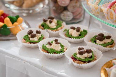 Beyaz bir masa üzerinde düzenlenmiş yeşil krema, çırpılmış krema ve çikolata şekerleriyle dolu canlı bir mini tart gösterisi, şenlikli durumlar veya tatlı masaları için idealdir.