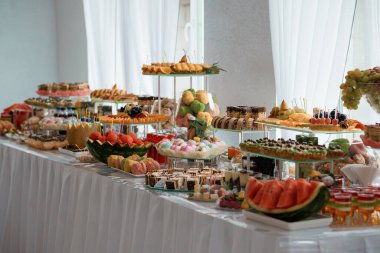 Renkli pastalar, meyve düzenlemeleri ve hamur işlerinin yer aldığı gösterişli bir tatlı büfesi beyaz kumaşlı ve doğal ışıklandırmalı parlak, modern bir iç mekan ortamında.