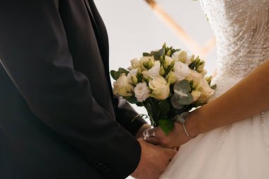 Gelin ve damadın bir buket beyaz gül ve yeşil okaliptüs ile el ele tutuşup, yumuşak ışıklandırma ve detaylı dantel elbiseyle samimi bir düğün anı sergiledikleri yakın plan.