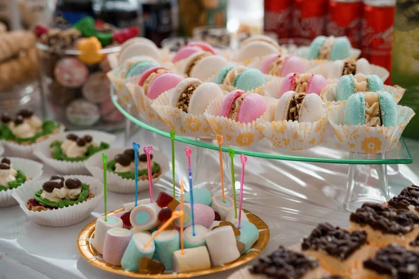 Pastel renkli badem kurabiyeleri, marşmelovlar ve çeşitli tatlılardan oluşan canlı bir çeşitlilik parlak ışıklandırma ve renkli dekorasyonlarla dolu bir parti ortamında katmanlı cam tezgahlarda düzenlenir.