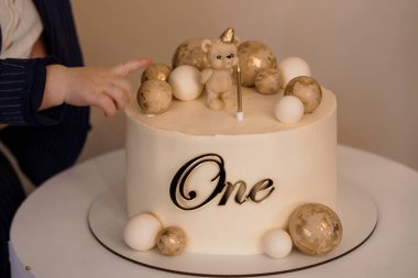 Bir çocuk, güzel dekore edilmiş ilk doğum günü pastasını, oyuncak ayıyla, altın aksanıyla ve yumuşak bej ve beyaz renk düzeniyle küresel dekorasyonlarla göstererek neşeli bir dönüm noktası yakalar.