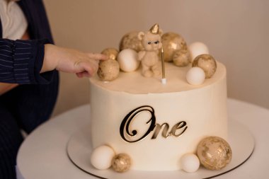 Bir çocuk, altın bir ayıcık, dekoratif yumurtalar ve neşeli bir dönüm noktasını yakalayan tek bir mumla süslenmiş krem rengi ilk doğum günü pastasına uzanır.