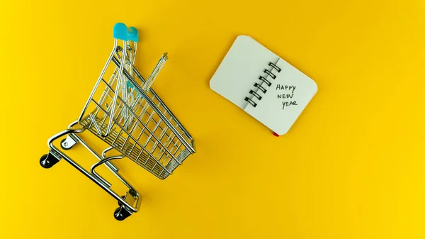 Sobrepasar el pequeño carrito de la compra y el cuaderno con el texto del Feliz Año Nuevo en él — Foto de Stock