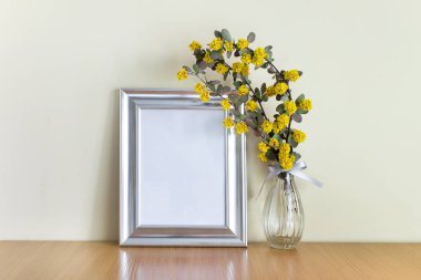 Boş gümüş çerçeve, cam vazoda sarı çiçekler tahta yüzeyde duruyor..