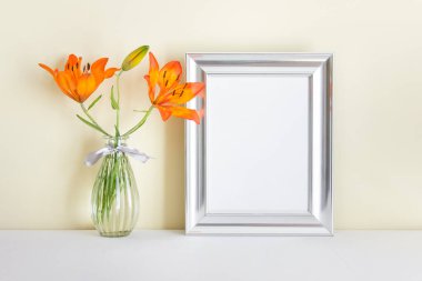 Boş uzay yüzeyi ve beyaz yüzey üzerinde duran cam vazoda turuncu zambak bulunan A4 boş gümüş çerçeve.