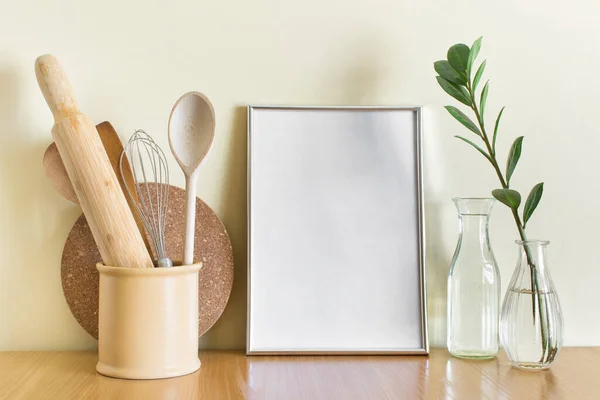 Gümüş metalik boş A4 çerçeve, mutfak ahşabı, cam vazolar ve yeşil bitki dalı olan model şablon..