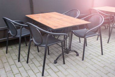 Kafenin dışında, ahşap masa ve siyah plastik sandalyeli..