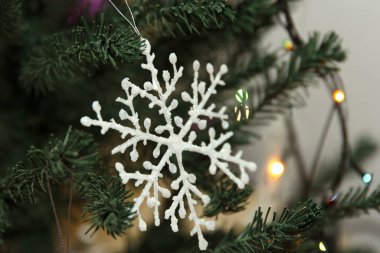 Yeşil bir Noel ağacına asılı beyaz bir kar tanesi süsü. Arkaplanda renkli ışıklar var, neşeli bir atmosfer yaratıyorlar.