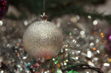 A close-up of a silver glittery Christmas ornament hanging among colorful tinsel. Background is blurred, creating a festive atmosphere