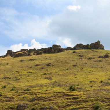 Kayalık bir yamaç, beyaz bulutlu mavi gökyüzünün zeminine doğru yükselir. Çimenler gür ve yeşildir. Gün ortasında doğal ortamda canlı bir kontrastlık yaratır. Güzel manzara.
