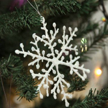 Yeşil bir Noel ağacına asılı beyaz bir kar tanesi süsü. Arkaplanda renkli ışıklar var, neşeli bir atmosfer yaratıyorlar.