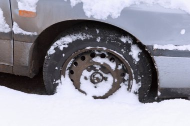 Bir araba lastiği kısmen kara gömülmüş. Tekerlek görünür deliklerle metaliktir. Kar zemini ve aracın alt kısmını kaplıyor.