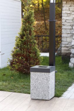 Modern, kare bir atık kutusu, desenli taş tabanlı ve siyah kapağı parktaki kaldırımlı bir geçitte duruyor. Yeşil çimenler, daima yeşil bir çalı ve sonbahar ormanı temiz, doğal bir ortam yaratır..